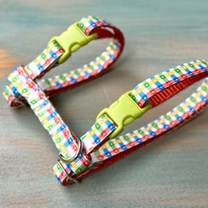 May include: A colorful pet harness with a plaid pattern in red, blue, yellow, and green. The harness has a red interior and lime green plastic buckles. The harness is designed for small pets.