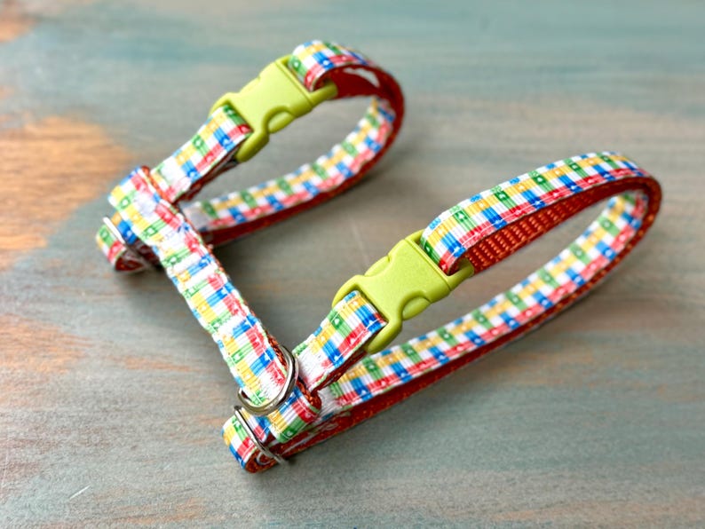 May include: A colorful pet harness with a plaid pattern in red, blue, yellow, and green. The harness has a red interior and lime green plastic buckles. The harness is designed for small pets.