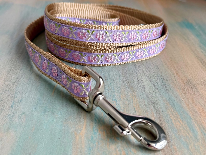 Puede incluir: Una correa de perro beige con un patr&oacute;n floral lavanda y un clip plateado.
