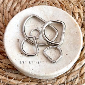 May include: A collection of silver-colored metal D-rings in various sizes, featuring smooth, rounded edges and a polished finish. The text 5/8", 3/4", and 1" is printed on the surface, indicating the specific measurements for these hardware components.