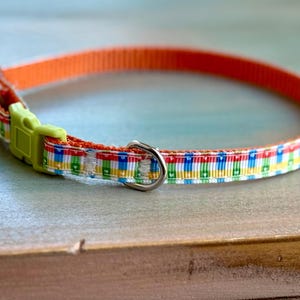 May include: A colorful pet collar with an orange nylon strap and a patterned ribbon. The ribbon features a repeating design of colorful blocks. The collar has a lime green plastic buckle and a silver metal D-ring.
