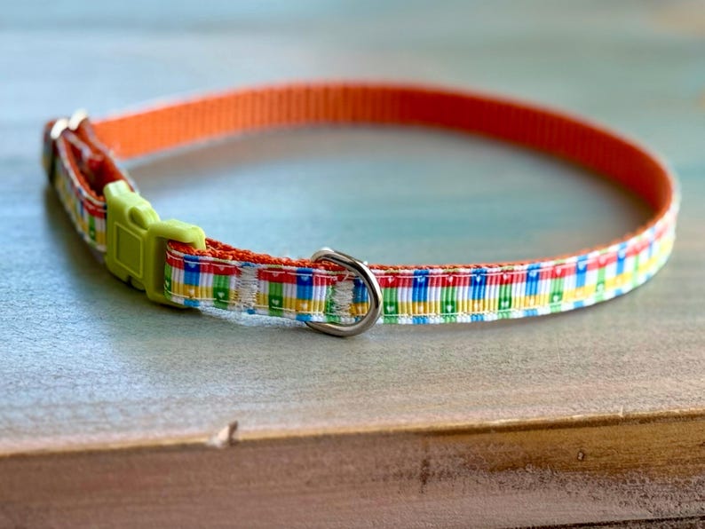 May include: A colorful pet collar with an orange nylon strap and a patterned ribbon. The ribbon features a repeating design of colorful blocks. The collar has a lime green plastic buckle and a silver metal D-ring.