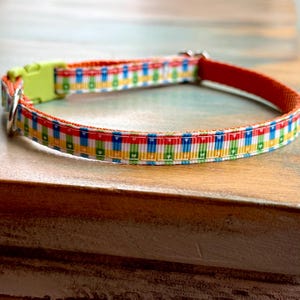 May include: A colorful dog collar with a repeating pattern of red, blue, yellow, and green rectangles. The collar has a lime green plastic buckle and a silver metal D-ring. The collar's interior is a solid red.