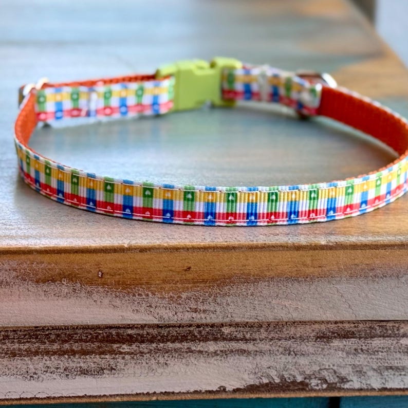May include: A colorful pet collar with a repeating pattern of red, blue, yellow, and green squares. The collar has an orange interior and a lime green plastic buckle. The collar is made of a durable, woven material.