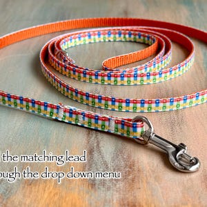May include: A colorful dog leash with a repeating pattern of red, yellow, green, and blue squares on a white background. The leash has an orange border and a silver metal clasp. Text on the image reads: "find the matching lead through the drop down menu."