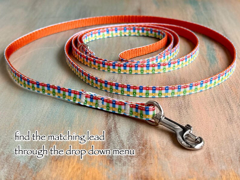 May include: A colorful dog leash with a repeating pattern of red, yellow, green, and blue squares on a white background. The leash has an orange border and a silver metal clasp. Text on the image reads: "find the matching lead through the drop down menu."