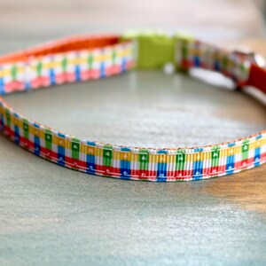 May include: A colorful dog collar with a repeating pattern of red, blue, green, and yellow squares. The collar has an orange interior and a lime green buckle. The collar is made of a durable material.