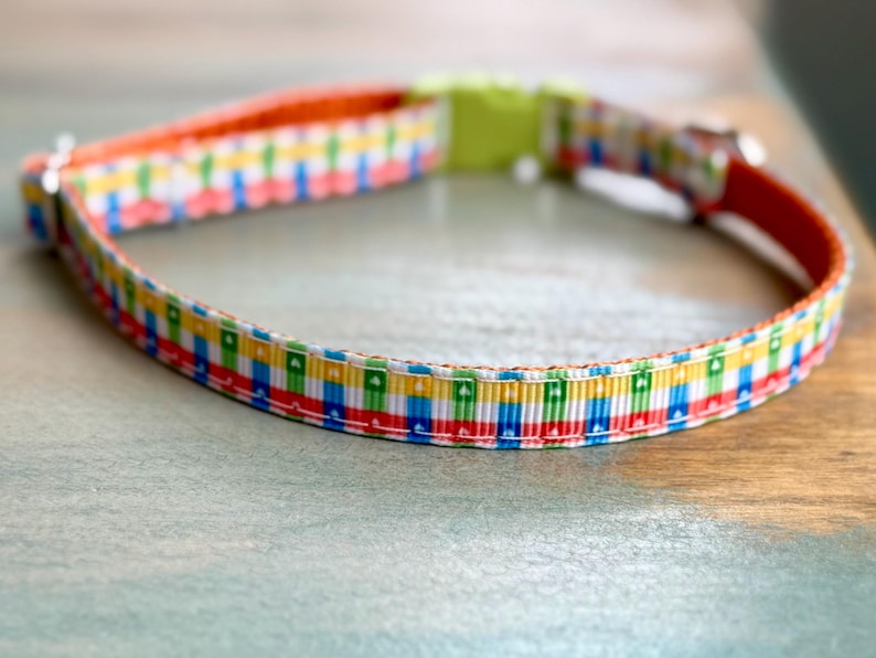 May include: A colorful dog collar with a repeating pattern of red, blue, green, and yellow squares. The collar has an orange interior and a lime green buckle. The collar is made of a durable material.