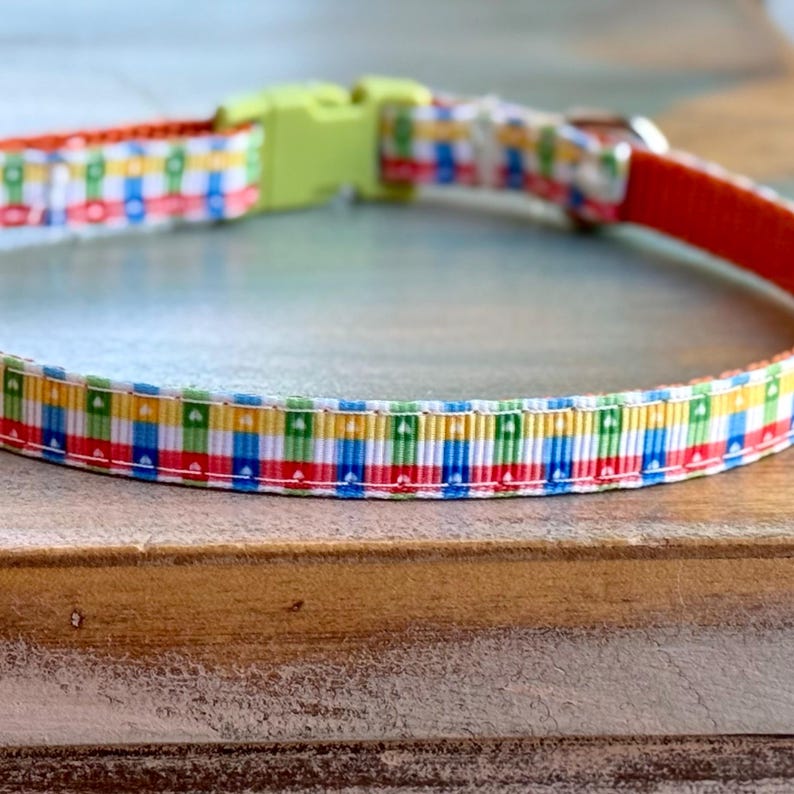 May include: A colorful dog collar with a repeating pattern of red, blue, yellow, and green blocks. The collar has a lime green plastic buckle and a red interior. The collar is designed for pets.