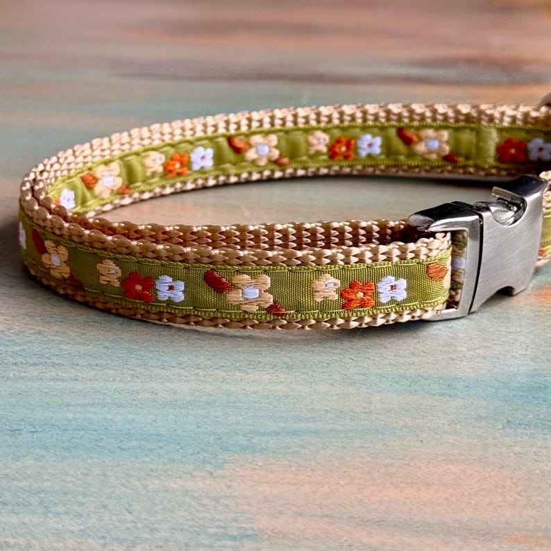 Op de afbeelding: Een close-up van een hondenhalsband met een bloemenpatroon. De halsband heeft een lichtgroen lint met geborduurde bloemen in oranje, wit en bruin. De randen zijn afgewerkt met een geweven beige materiaal en heeft een zilveren metalen sluiting.