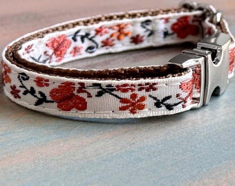 May include: A pet collar featuring a white fabric strap with an embroidered floral pattern in shades of red, orange, and black. The collar includes a brown nylon backing and a polished silver-toned metal side-release buckle for secure fastening.