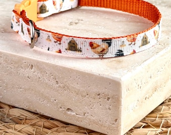 Puede incluir: Un collar para mascota con una cinta blanca que presenta un patrón repetitivo de pollos y pequeños gallineros de madera. El collar tiene un reverso de nailon naranja sólido, una hebilla de plástico naranja claro y una anilla en D de metal plateado.