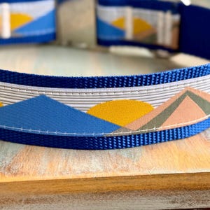 May include: A blue dog collar with a white and gray geometric pattern featuring a yellow sun and blue mountains.