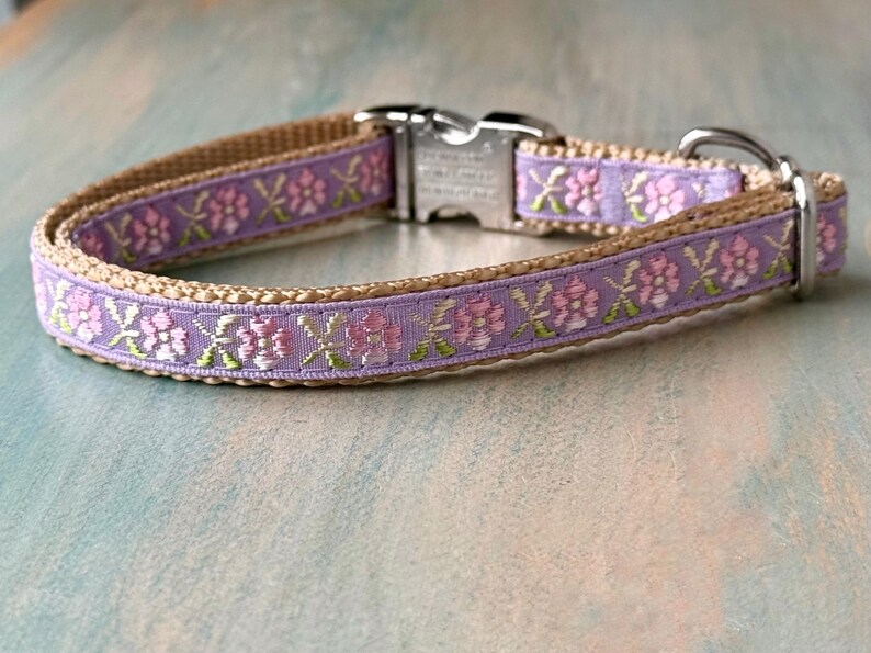 Puede incluir: Un collar para perro de color marr&oacute;n claro con un patr&oacute;n floral morado y rosa. El collar tiene una hebilla plateada y un anillo en D plateado.