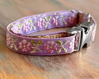 May include: A lavender fabric collar featuring an embroidered floral pattern in shades of pink, light green, and gold. The collar includes a polished silver-toned metal side-release buckle and an adjustable slider for securing the strap.