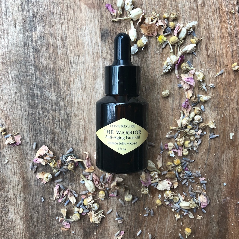 May include: A dark brown glass bottle with a black dropper top and a gold label that reads "LOVERDURE THE WARRIOR Anti-Aging Face Oil Immortelle + Rose 1 fl oz". The bottle is surrounded by dried flower petals on a wooden surface.