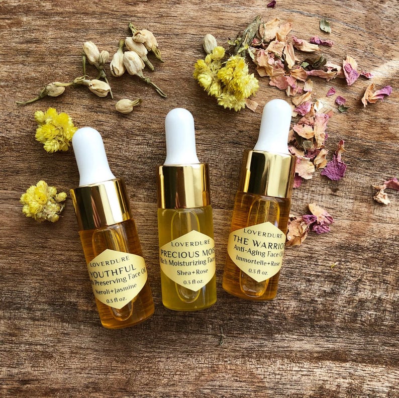May include: Three small glass bottles with gold caps and labels. The bottles contain face oils with the following names and ingredients: Youthful (Neroli + Jasmine), Precious Moisturizing (Shea + Rose), and The Warrior (Immortelle + Rose). Each bottle is 0.3 fluid ounces.
