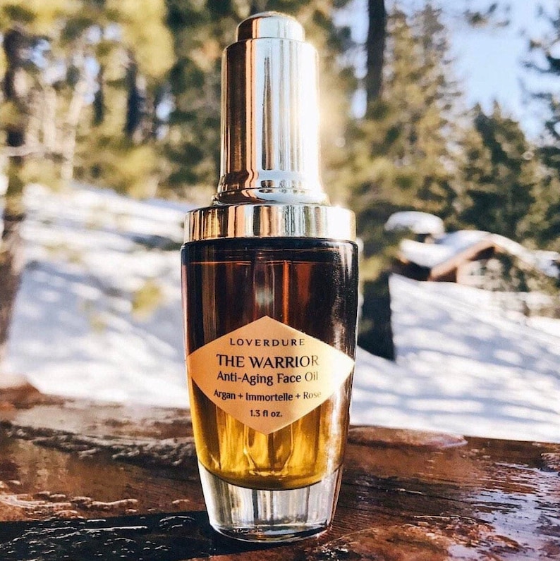 May include: A glass bottle of LOVERDURE THE WARRIOR Anti-Aging Face Oil with a gold dropper and cap. The bottle is filled with a golden oil and the label is white with black text. The bottle is sitting on a wooden surface.