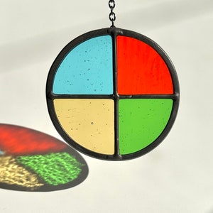 May include: A round stained glass suncatcher with a black metal frame. The glass is divided into four sections: blue, red, yellow, and green. A black chain is attached for hanging. The suncatcher casts a colorful shadow.