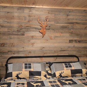 May include: A bedroom featuring a wall covered in horizontal gray wooden planks with a mounted wooden deer head silhouette. A black metal bed frame holds pillows and a quilt patterned with blue, tan, and black plaid squares and deer motifs.