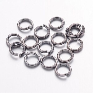 Könnte beinhalten: Eine Gruppe von 15 schwarzen Sprungringen, eine Art von Schmuckzubehör, liegt auf einer weißen Oberfläche verstreut. Die Ringe sind aus Metall und haben eine glatte, glänzende Oberfläche.