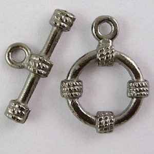 Könnte beinhalten: Zwei silberfarbene Metallverschlüsse zum Schmuckmachen. Ein Verschluss ist ein Toggle-Verschluss mit einer runden Schlaufe und einer Stange mit einem dekorativen Ende. Der andere Verschluss ist ein runder Ring mit vier dekorativen Akzenten.