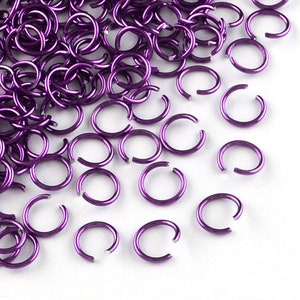May include: A large pile of purple jump rings, scattered on a white surface. The jump rings are small, round, and made of metal.