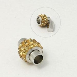 Könnte beinhalten: Ein goldfarbener Magnetverschluss mit einer runden, mit klaren Kristallen besetzten Oberfläche. Der Verschluss ist aus Metall und hat eine polierte Oberfläche.
