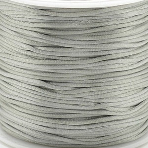 May include: A spool of light gray satin cord. The cord is tightly wound, revealing a smooth, reflective surface. The color is a neutral, versatile shade, suitable for various crafting projects.