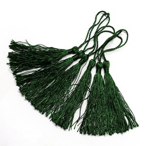 Puede incluir: Un conjunto de siete borlas de color verde oscuro con hebras largas y delgadas. Las borlas están atadas en la parte superior con un cordón delgado.