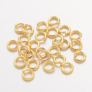 Könnte beinhalten: Eine Ansammlung kleiner, goldfarbener Sprungringe, die auf einer weißen Oberfläche verstreut sind. Die Ringe sind offen und bereit zum Verbinden. Diese Schmuckzubehörteile sind ideal für Bastelarbeiten und die Schmuckherstellung.