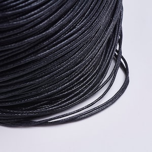 May include: A close-up of a spool of black waxed cotton cord. The cord is tightly wound and has a smooth, polished finish.