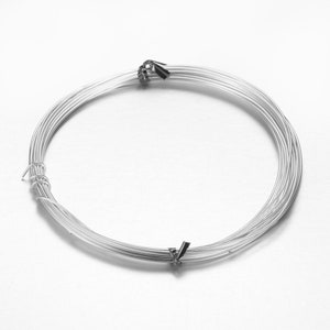 Peut inclure: Une bobine de fil métallique argenté, enroulée en cercle et fixée avec une attache métallique. Le fil semble lisse et souple, idéal pour la création de bijoux ou d'artisanat. L'image est une vue rapprochée, mettant en évidence la texture et la forme du fil.