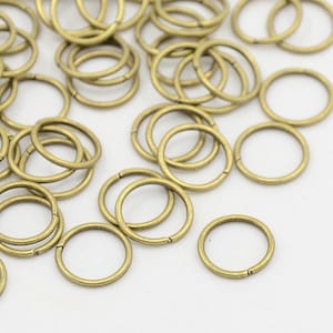 Op de afbeelding: Een verzameling kleine, messingkleurige metalen ringen, sommige open en sommige gesloten, verspreid over een wit oppervlak. De ringen zijn cirkelvormig en lijken van metaal te zijn. De afbeelding is een close-up.