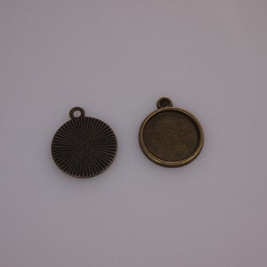 May include: Two bronze-colored round pendant blanks. One has a ridged, sunburst design on the front. The other has a flat, recessed area for inserting a design, with a loop at the top for a chain.