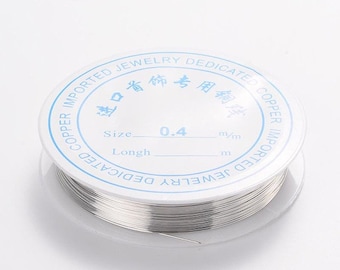Puede incluir: Una bobina de alambre de color plateado con una etiqueta blanca que dice "IMPORTED JEWELRY DEDICATED COPPER IMPORTED COPPER JEWELRY DEDICATED" y "Size 0.4 mm/m Longth m".