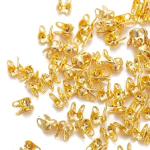 Könnte beinhalten: Eine große Gruppe goldfarbener Metall-Quetschperlen. Die Perlen haben an jeder Seite eine kleine Schlaufe und eine abgerundete Oberseite.