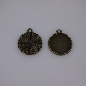 May include: Two antique bronze-colored round pendant trays. One pendant has a textured sunburst design, and the other is a plain bezel. Both have a small loop at the top for attaching to a chain or cord. These are ideal for jewelry making.