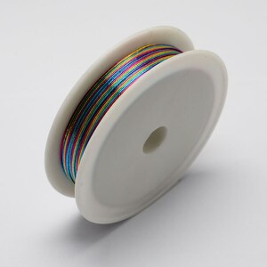 Op de afbeelding: Een spoel met iriserende metallic draad in een regenboog van kleuren, waaronder rood, oranje, geel, groen, blauw en paars. De draad is gewikkeld om een witte plastic spoel.