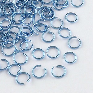 Op de afbeelding: Een grote hoeveelheid kleine, lichtblauwe metalen springringen. De ringen zijn allemaal even groot en liggen verspreid op een wit oppervlak.