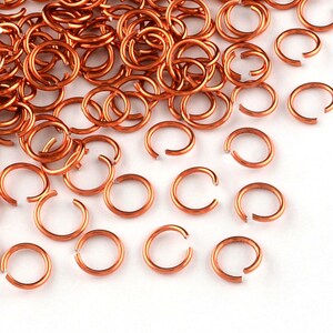 Op de afbeelding: Een grote stapel koperkleurige springringen, met een paar verspreid over een wit oppervlak. De springringen zijn klein en rond, met een diameter van ongeveer 6 mm.