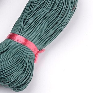 May include: A close-up of a teal-coloured cord wound into a spool, held together with a pink band. The cord has a slightly textured surface and is set against a white background. Ideal for crafts and various projects.