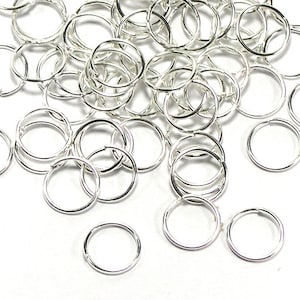 Könnte beinhalten: Eine Ansammlung kleiner, glänzender, silberfarbener Biegeringe, die auf einer weißen Oberfläche verstreut sind. Die Ringe sind kreisförmig mit einer kleinen Öffnung, was darauf hindeutet, dass sie für die Schmuckherstellung oder Bastelarbeiten bestimmt sind.