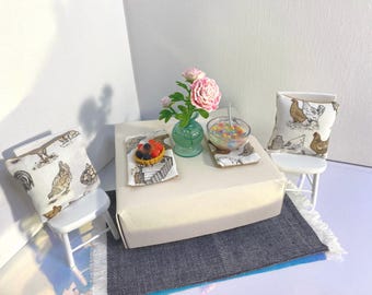 May include: Miniature dining scene with a white table, two small chairs, and decorative pillows. A vase of pink flowers, a bowl of colorful candies, and a fruit tart are on the table. The pillows and placemats feature a chicken motif. A blue and gray rug is on the floor.