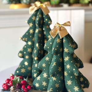 May include: Two green fabric Christmas trees with gold snowflake and star patterns, topped with gold bows. The trees sit on a wooden base, with red berries and pinecones in front. Other similar trees are in the background.