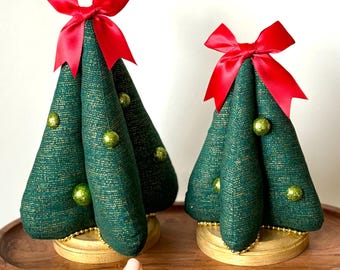 May include: Two decorative Christmas trees made of green fabric with gold accents, topped with red bows and adorned with green glitter ornaments. The trees sit on wooden bases and are displayed on a wooden tray.