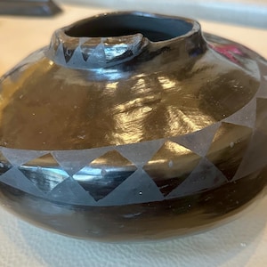 May include: A black ceramic vase with a unique shape and a glossy finish. The vase features a geometric diamond pattern around the body and a decorative rim. The pottery is handmade and has a rustic, artisanal feel.