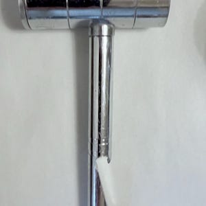 May include: A shiny, chrome-colored tool with a T-shaped design. The top section is cylindrical with horizontal grooves, and the vertical section has a ridged design. A small piece of white material is attached to the bottom.