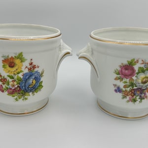 May include: Two white ceramic planters with gold trim and handles. Each planter features a floral design with colorful flowers. One has a blue rose, the other has a pink rose.