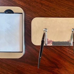 May include: A gold-toned desk set featuring a rectangular notepad holder and a pen holder with two pens. The notepad holder has a black accent at the top. The pens are black with silver accents. The set is on a dark wood surface.